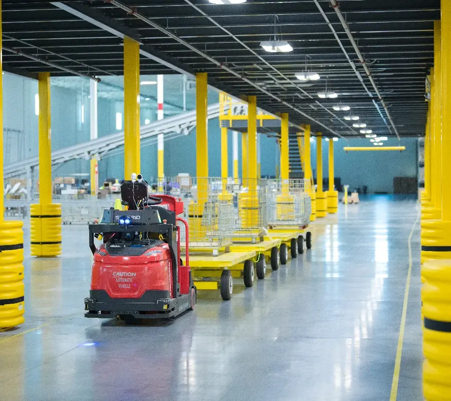 An automated guided vehicle towing several carts through a modern warehouse with yellow safety pillars.