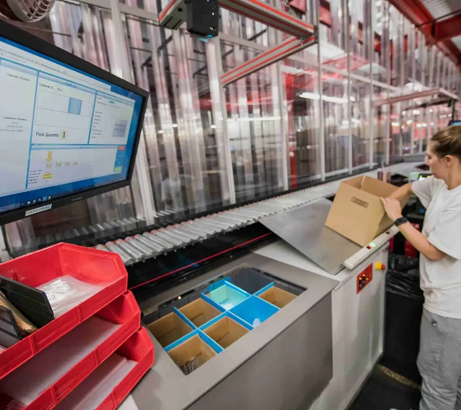 Warehouse worker at a goods-to-person station fulfilling orders using an automated conveyor system.