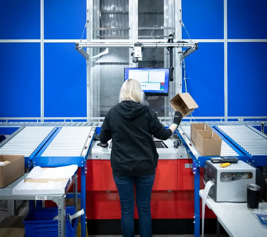 A warehouse operator packs cardboard boxes at a modern goods-to-person fulfillment station.