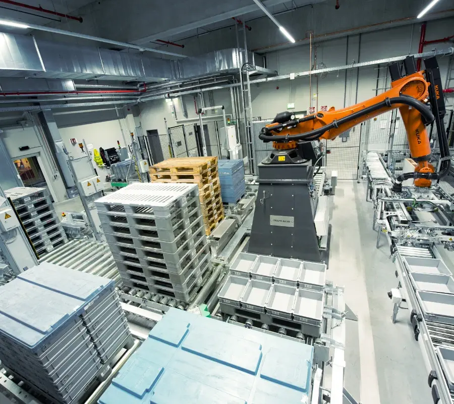 An automated robot arm handles crates and pallets on a conveyor system in an industrial warehouse.