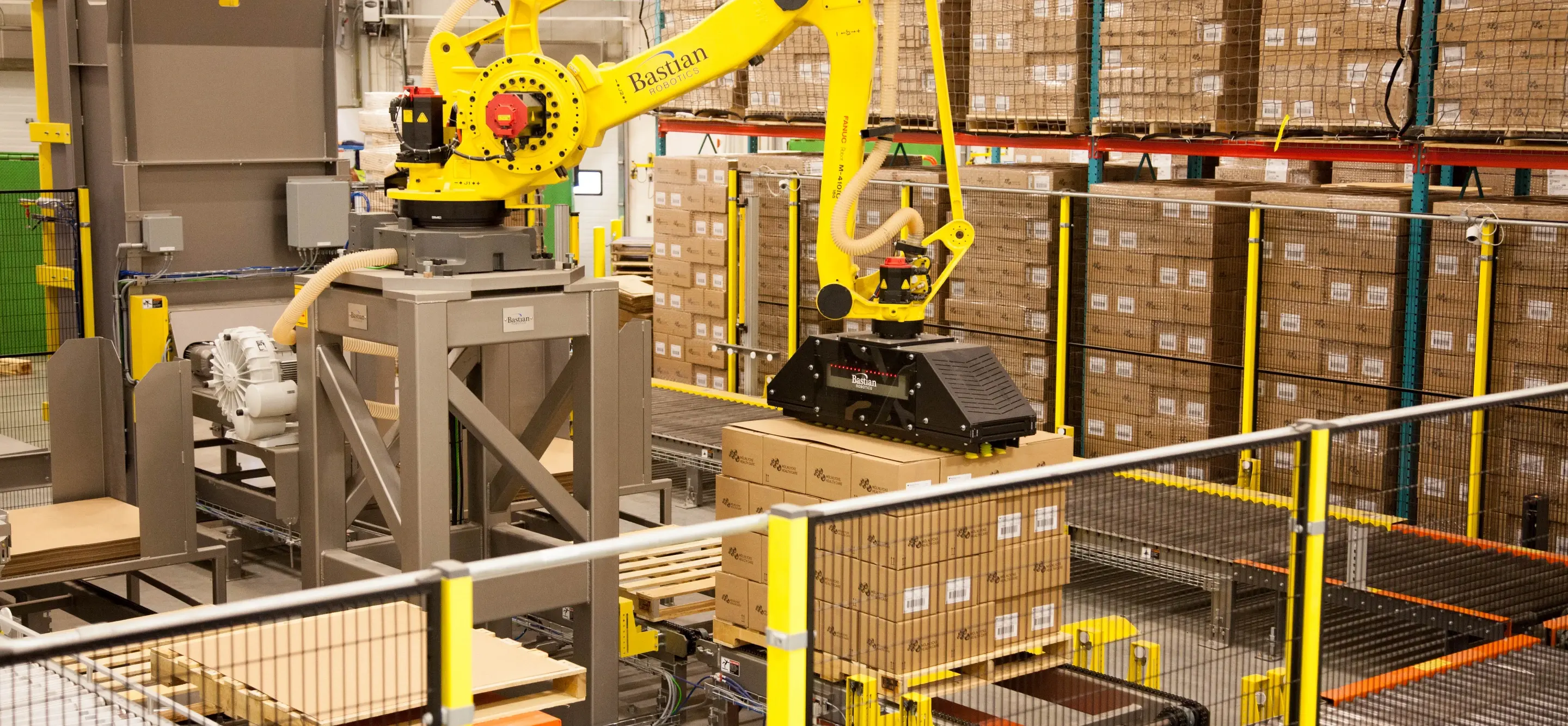 Yellow industrial robotic arm palletizing boxes onto a wooden pallet in a warehouse facility.
